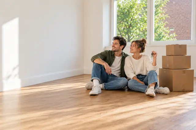A smiling couple sitting on the floor of an empty, clean flat beside moving boxes, one holding the keys